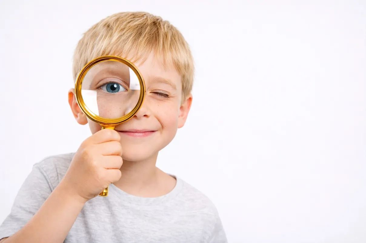 Bambino biondo sorride e guarda in camera tenendo una lente d’ingrandimento davanti all’occhio destro, che appare ingrandito.