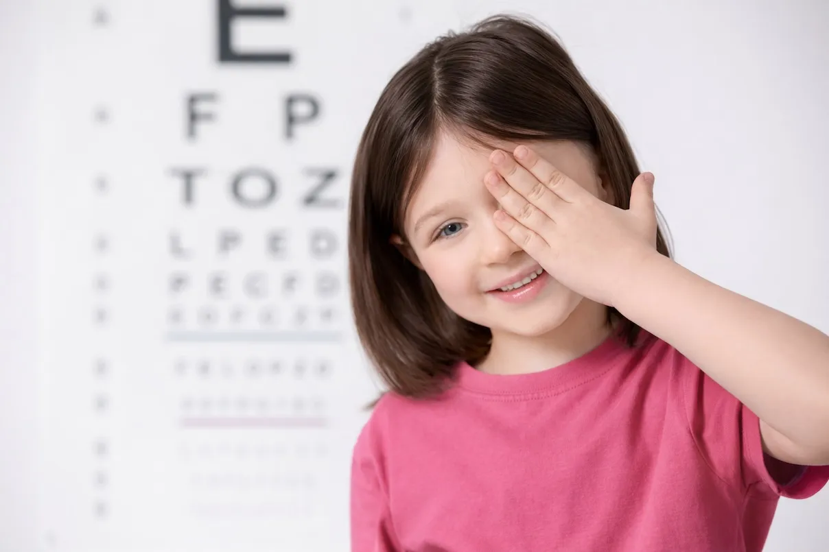 Bambina sorridente con capelli castano scuro che copre l’occhio destro con la mano durante un esame della vista; indossa una maglia rosa e davanti a lei si vede sfocata una tabella ottometrica con lettere nere.
