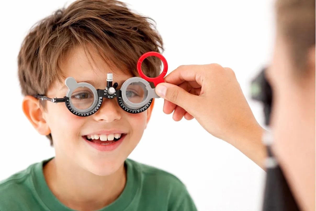Bambino sorridente con capelli castani che indossa una montatura da prova per la vista; un adulto gli avvicina una lente rossa davanti all’occhio destro durante un esame oculistico su sfondo bianco.